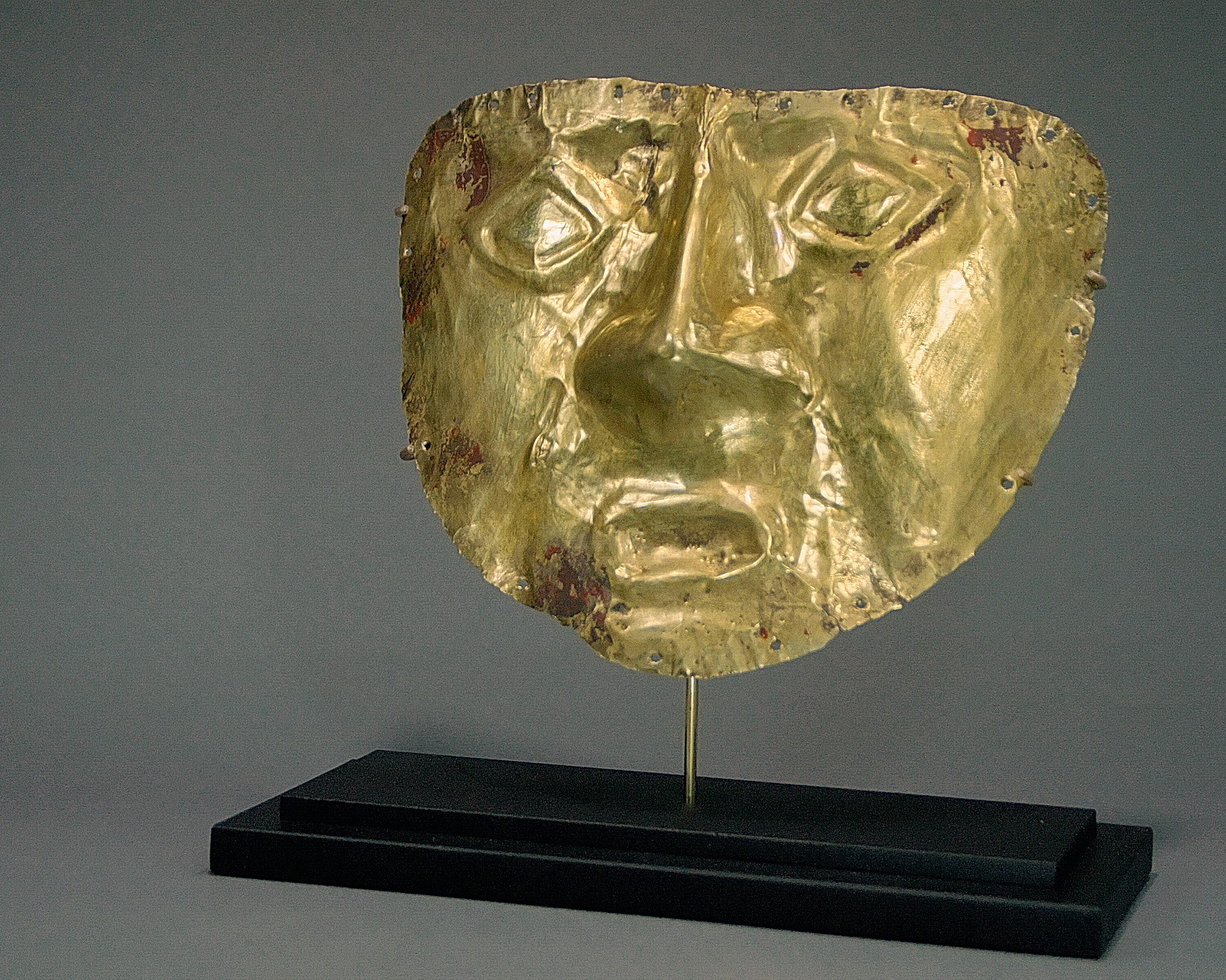 Peru, Inca gold Mask with Traces of the Original Cinnabar Patina
Inca Gold mask in one beaten sheet of gold with the nose protruding and has diamond shaped eyes.  Earlier masks had noses which were made by hammering the gold  on a wooden form while later masks were made by adding a separate nose piece and stapling it with gold strips to a cutout for the nose.  There are 20 punched holes around the parameter of the mask which was probably were made to sew the mask to a mummy bundle for the afterlife as opposed to use it for ceremonial use.   Similar masks are illustrated in OroDel Antiguo Peru, plates 203 & 204.   Collected prior to 1970 by the former president of Playtex.
Media: Metal
Dimensions: Height : 6 1/2"x Width: 7 3/4"     Weight 54 grams
Price Upon Request
n4035