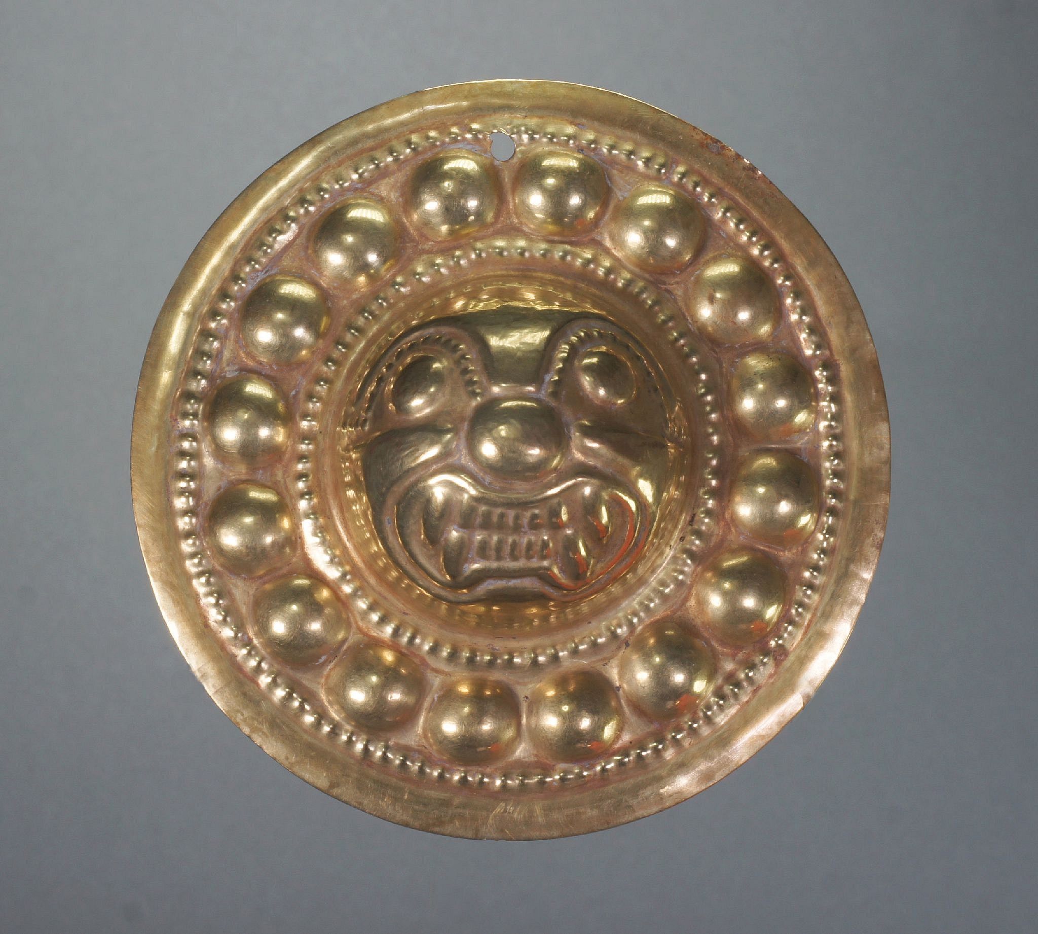 Ecuador, Narino Gold Embossed Ornament Decorated with a Feline Head
These ornaments have a suspension on the rim and were either ear ornaments or pendants.  The designs vary, from concentric circles to heads in high relief, ranging from human to animal faces.  The feline face is the largest of the head types.  Similar examples are illustrated in El Dorado by Warwick Bray, p.213.  Ex. New York collector, prior to 1970.
Media: Metal
Dimensions: Diameter: 3.9"   Weight: 26.7gramsGold alloy:  Gold 73.4%, Silver 17.9%, Copper 8.3% and Sn. Tin .01%
&bull;SOLD
N7060