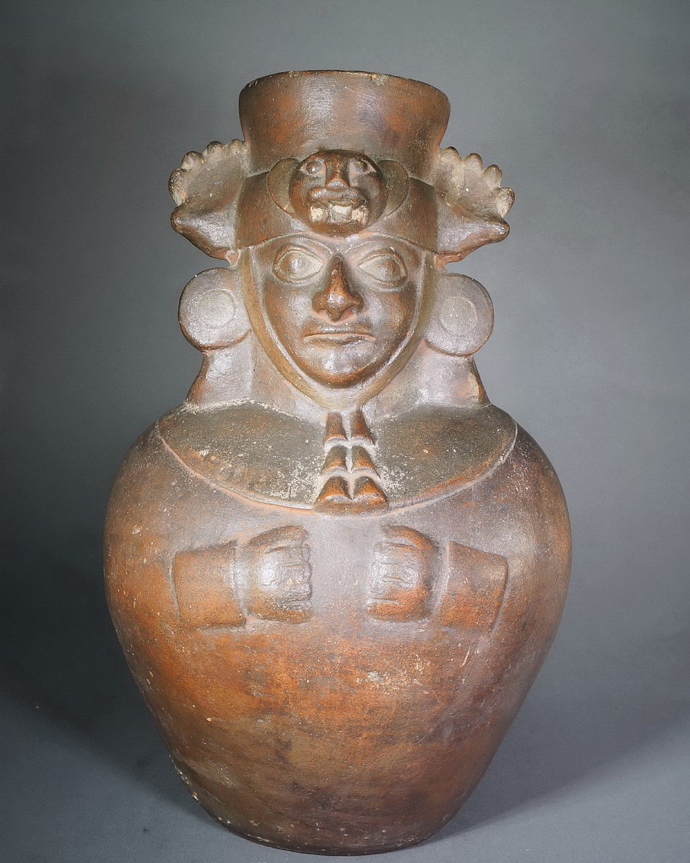 Peru, Moche III/IV Redware Mold Made Jar Depicting a Dignitary
The figure is wearing a head cloth underneath a Puma head ring with straps ending in tassels and large paws, with its polka-dot tail down his back.   He also wears large ear spools which denote his status.  He holds his hands to his chest below a semi-circular breast ornament.  These vessels were made in a two-part mold, one for the front and the other for the back.
Media: Ceramic
Dimensions: Height: 12 1/2"
Price Upon Request
n6049
