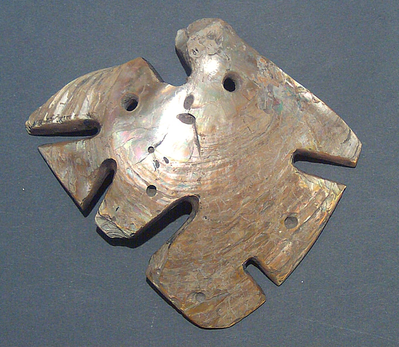 Ecuador, Mother of Pearl Fish Ornament
This shell was carved to represent a highly abstracted fish.  Drill holes indicate that it was worn as a pendant.  Ancient Ecuadorean coastal societies often depicted local fauna, both naturalistically and as abstract shapes.  Shells, which grew in abundance in the warm coastal waters off Ecuador, were coveted as prestigious items throughout the Andes.  A similar example is illustrated in Francisco Valdez and Diego Veintimilla’s Amerindian Signs: 5,000 Years of Pre-Columbian Art in Ecuador (1992: fig. 63).
Media: Shell
Dimensions: Width: 4 1/4" x Height: 3 3/4"
&bull;SOLD
94302b