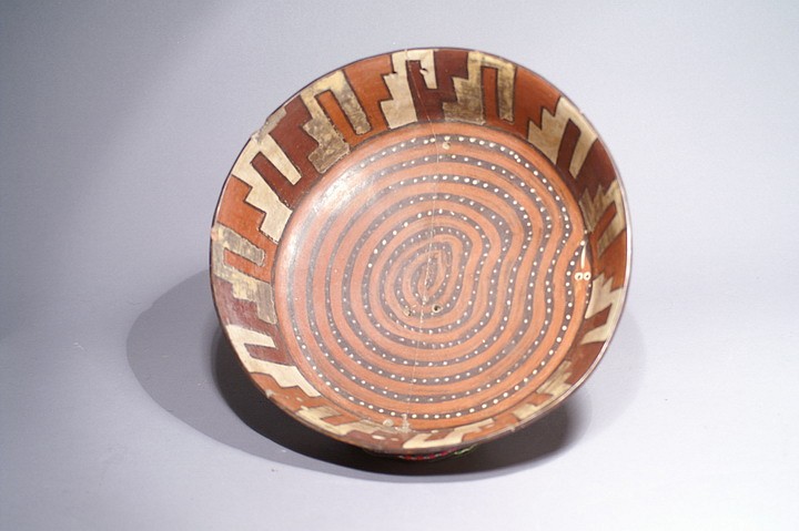 Peru, Nazca Polychrome Dish with Serpent Motif
This wonderful Nazca ceramic dish is painted with a spiral serpent motif at the bottom.  The serpent is painted with a pattern of white dots.  The inner edge of the dishâ€™s high wall is painted with a step-fret pattern in alternating in colors of maroon, white and orange.  The step-fret motif likely represents stairs to a temple or stepped agricultural plains, both of which were important to the Nazca.  On the outside rim of the dish are 10 painted zoomorphic faces with long noses.  The bottom of the dish is painted black.   There is evidence of ancient repairs made to the dish, which can be seen via small drill holes made on each side of a crack and binding applied to the crack. Benno Mattel collection, Punta del Este, Uruguay.
Media: Ceramic
Dimensions: Diameter 9 1/2" x height 2 1/2"
Price Upon Request
N1032